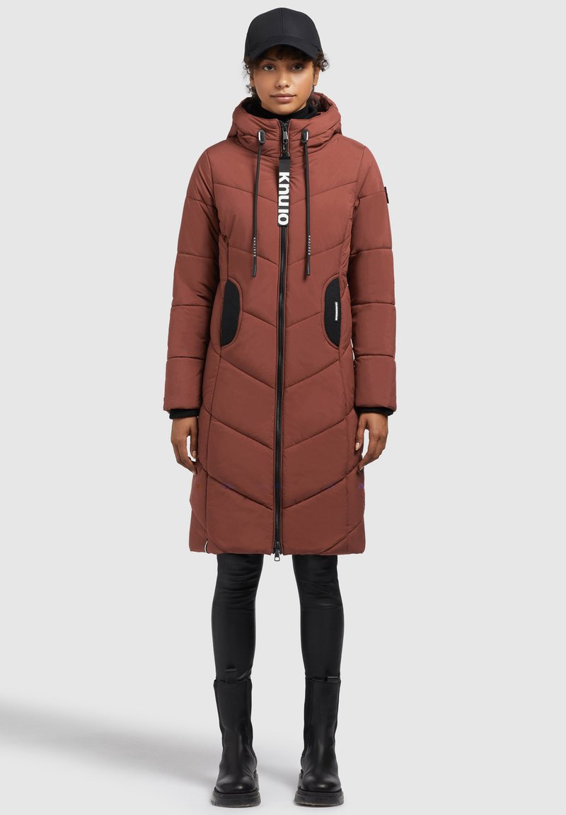 Brun polstret lang frakke med chevronmønster, hætte, frontlynlås, sorte detaljer og to sidelommer. Båret over tætsiddende sorte bukser og støvler.