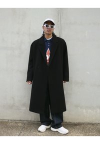 Manteau noir long avec un design structuré, porté sur un polo bleu foncé avec un motif en losange rouge, complété par des baskets blanches et des lunettes de soleil.