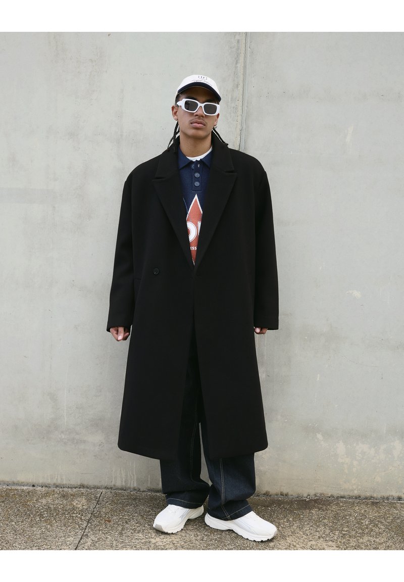 Manteau noir long avec un design structuré, porté sur un polo bleu foncé avec un motif en losange rouge, complété par des baskets blanches et des lunettes de soleil.