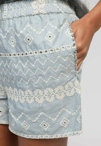 Main reposée dans la poche d'un short bleu clair avec des motifs géométriques brodés en blanc et de petits strass le long du bord de la poche.