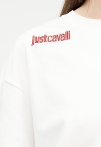 Λευκό φούτερ με κόκκινη κεντημένη επιγραφή "Just Cavalli" κοντά στον ώμο, φοράει ένα άτομο με μακριά καστανά μαλλιά που είναι ορατά από τη μία πλευρά.