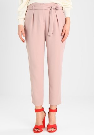 Frau trägt rosafarbene, hoch taillierte, schmal zulaufende Hose mit gebundenem Gürtel, kombiniert mit leuchtend roten Sandalen mit Absatz, steht vor weißem Hintergrund.