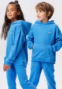 Twee kinderen in bijpassende blauwe hoodies en joggingsbroeken, zij aan zij staand tegen een witte achtergrond.