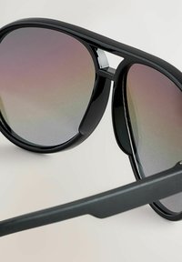 Lunettes de soleil aviateur noires avec des verres dégradés, présentant une monture en plastique épurée et un détail découpé distinctif sur le front.