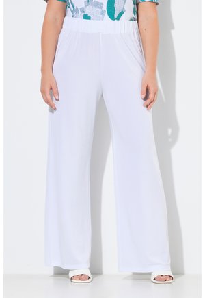 Kobieta ubrana w luźne białe spodnie z elastycznym pasem i bocznymi kieszeniami, w zestawie z białymi sandałami na otwartym palcu, stoi na tle białego tła.