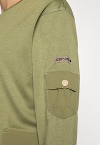 Oliven grønn sweatshirt med teksturert stoff, med en lomme på ermet og en logoetikett. Enkel, funksjonell design.