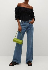 Schwarzes schulterfreies Oberteil aus strukturiertem Stoff, hellblaue hochgeschnittene weite Jeans, und eine glänzende grüne zylindrische Handtasche. Schwarze Sandalen.