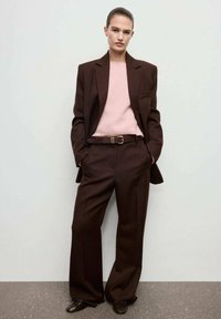 Donna in piedi con le mani nelle tasche, che indossa un completo marrone con pantaloni a gamba larga, un maglione rosa e scarpe marroni, di fronte a un muro bianco.
