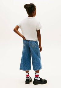 Biała koszulka z krótkim rękawem, szerokie niebieskie dżinsowe kuloty, paskowane skarpetki w czerwono-biało-niebieskie paski, czarne loafersy, jednolite tło.