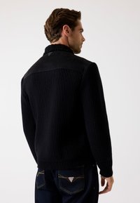 Schwarzer Strickpullover mit einer strukturierten, gewebten Oberfläche und Wildlederakzenten an den Schultern. Verfügt über einen hohen Kragen und gerippte Manschetten.