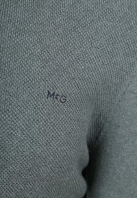 Grauer Strickpullover mit einem strukturierten Muster und einem kleinen gestickten Logo "M&G" am Ärmel. Weicher Stoff mit einer figurbetonten Passform.