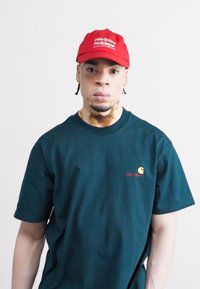 Căciulă roșie cu textul "Sărbători Hardcore", tricou bleumarin cu un logo mic galben și roșu. Design simplu, mâneci scurte și o croială lejeră.