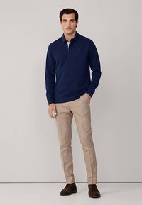 Marinblå stickad pikétröja med lång ärm och krage, kombinerad med ljusbeiga skräddarsydda byxor och mörkbruna skor. Enkel design utan mönster.