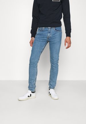 Personne portant un jean slim bleu clair, des baskets blanches avec des accents noirs et une chemise noire à manches longues, debout devant un fond uni.