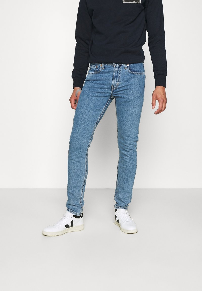 Personne portant un jean slim bleu clair, des baskets blanches avec des accents noirs et une chemise noire à manches longues, debout devant un fond uni.