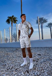 Camisa blanca de manga corta y pantalones cortos a juego con patrones de hojas tropicales marrones. Combina con zapatillas blancas y calcetines; tejido suave y ligero.