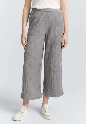 Kvinde iført løstsiddende grå krøllet viddebenbukser med elastisk talje og hvide sneakers, stående mod en ensfarvet baggrund.