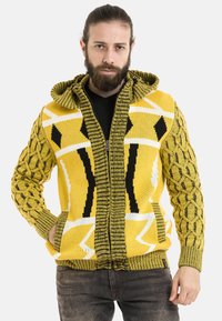 Gelbe Strickjacke mit Kapuze; verfügt über texturierte Ärmel, geometrische Muster in Schwarz und Weiß sowie Fronttaschen.