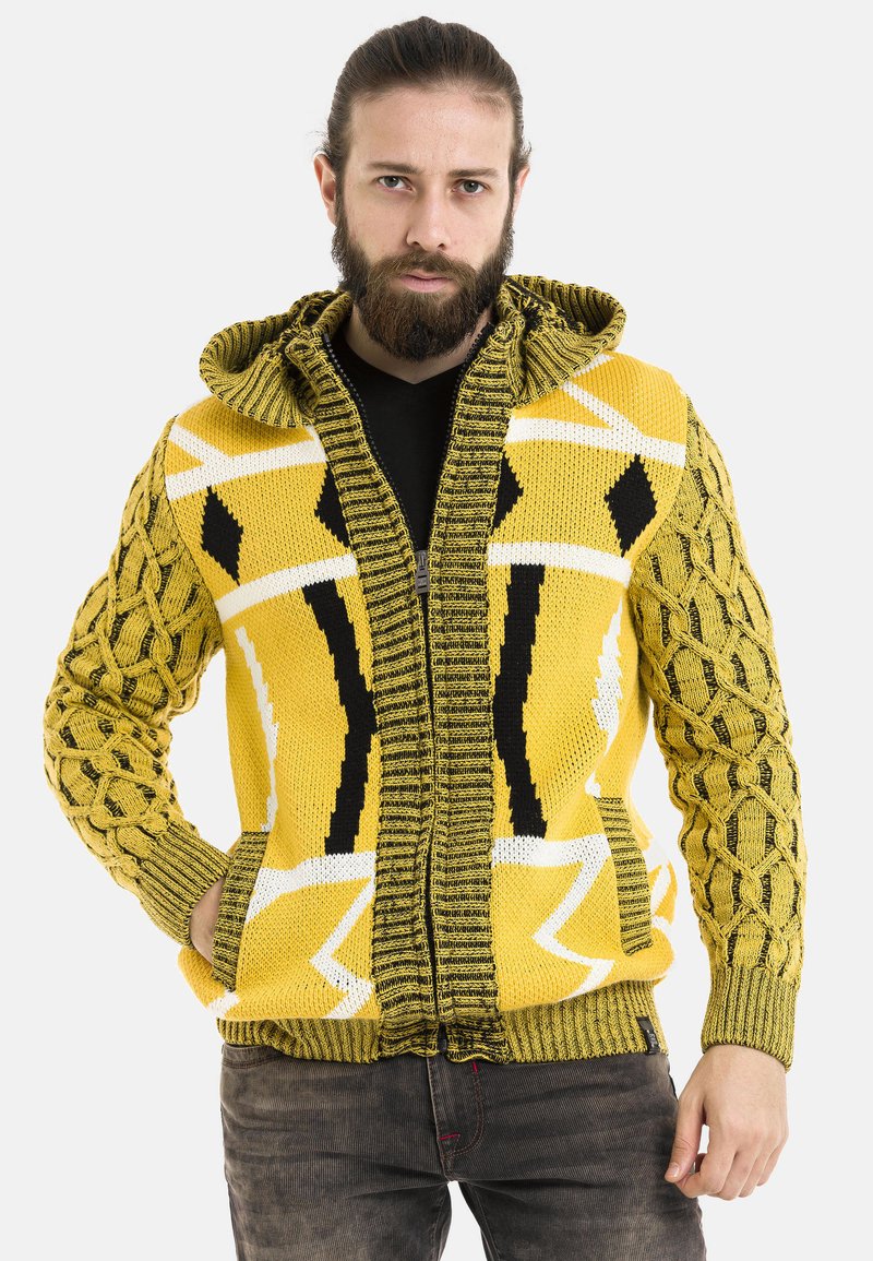 Gelbe Strickjacke mit Kapuze; verfügt über texturierte Ärmel, geometrische Muster in Schwarz und Weiß sowie Fronttaschen.