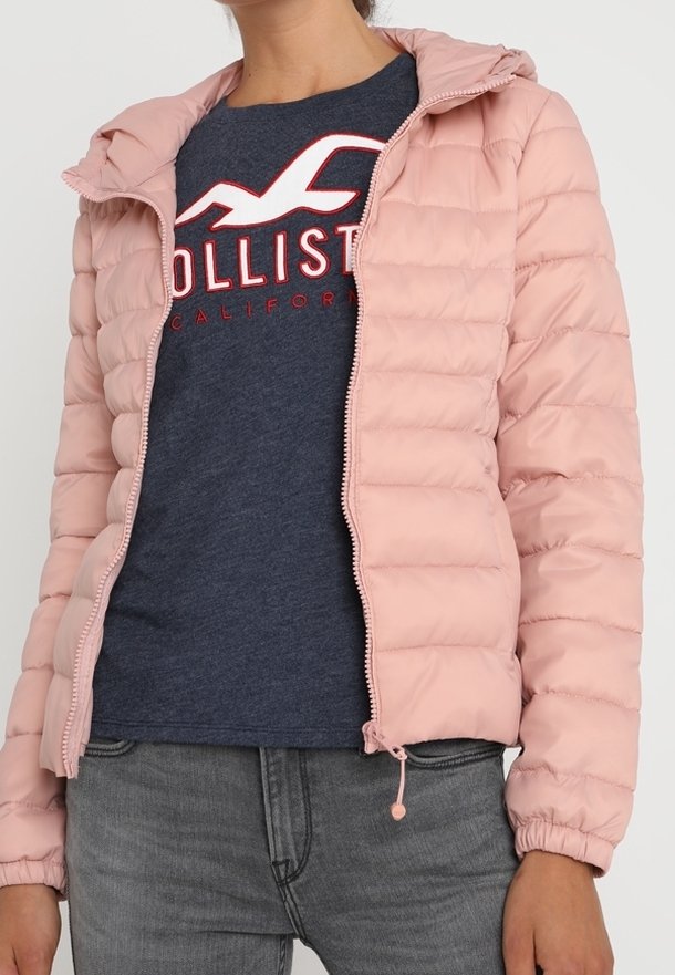 Light pink puffer jacket with a hood, zip closure, elastic cuffs, and drawstring hem. Worn over a dark blue shirt with red text.