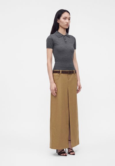 Femme debout portant un polo gris ajusté à manches courtes, une longue jupe beige fendue devant, une ceinture marron et des talons noirs ouverts sur fond blanc uni.