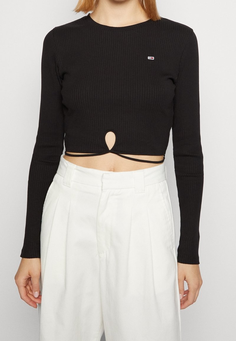 Donna che indossa un crop top nero a coste con scollatura a goccia e pantaloni bianchi a vita alta plissettati su sfondo bianco.