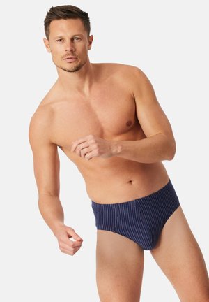 Uomo senza maglietta con capelli castani corti che indossa slip a righe blu navy posa contro uno sfondo bianco.