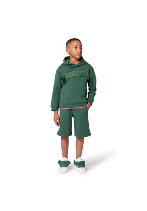 Sweat-shirt à capuche verte avec des détails tressés, associé à un short assorti. Baskets blanches remarquables avec des accents verts. Texture lisse, design décontracté.