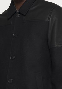 Svart ullblandad jacka med svarta läderaxelpaneler, fyra knappar och sidofickor. Har en klassisk krage och skräddarsydd design.