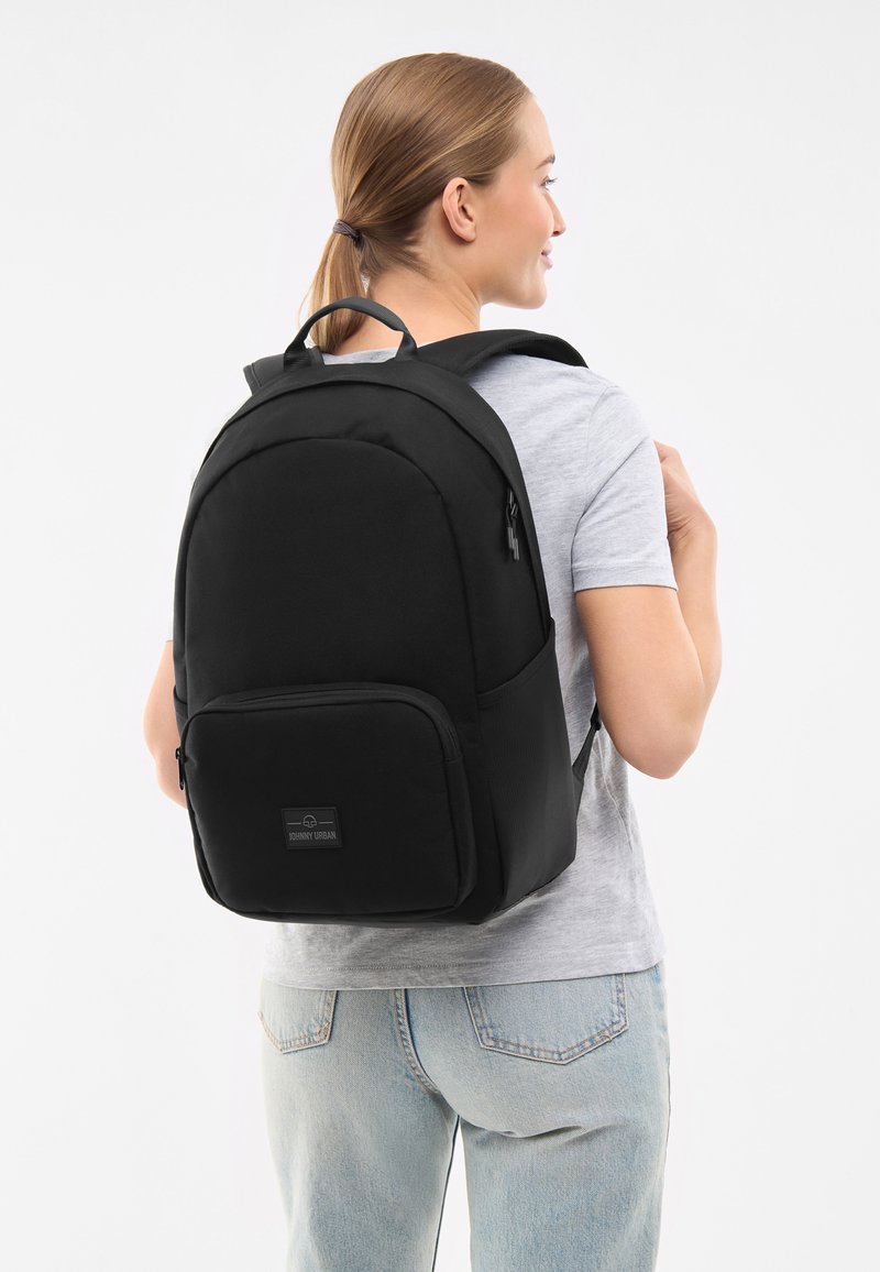 Svart ryggsäck tillverkad av hållbart tyg, med en framficka med dragkedja, vadderade axelremmar och ett handtag på toppen för bärande.