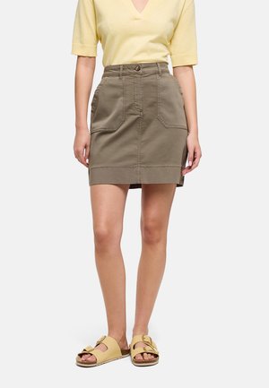 Mujer con top amarillo de manga corta, falda de talle alto color verde oliva con bolsillos frontales y sandalias beige con hebilla, de pie sobre fondo blanco.