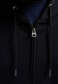 Felpa nera con zip e coulisse. Tessuto morbido e interni texturizzati. Presenta una maglietta blu navy in contrasto sotto.