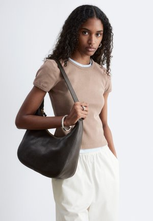 Joven con el pelo largo y rizado, vistiendo una camiseta beige ajustada y pantalones blancos, sosteniendo un gran bolso de hombro de cuero marrón oscuro.