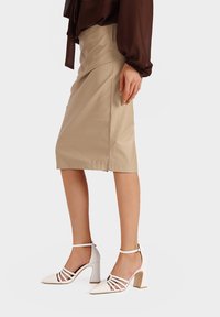 Gonna a matita in pelle beige abbinata a sandali bianchi con tacco, dotati di una cinghia alla caviglia e design a incrocio, su uno sfondo neutro.