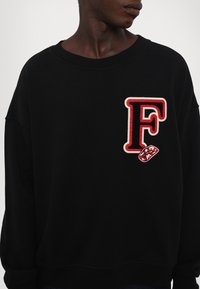Zwarte sweatshirt met een grote rode "F" geborduurd op de voorkant, vergezeld van een klein lippenstiftpatroon. Zachte stof, ontspannen pasvorm.
