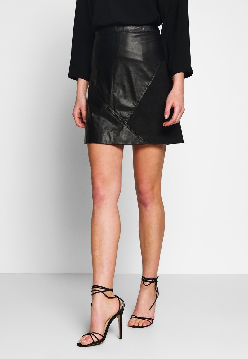 Svart läder mini kjol med diagonala sömmar, som har en figurnära midja och slät yta. Kombinerad med svarta högklackade sandaler med remmar.