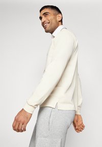 Long-sleeve cream shirt with side slits, paired with light gray sweatpants featuring a logo. Fabric appears soft with a relaxed fit.
