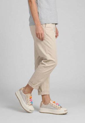 Personne portant un pantalon beige et des baskets claires avec des breloques en plastique multicolores sur les lacets, debout sur un sol gris.