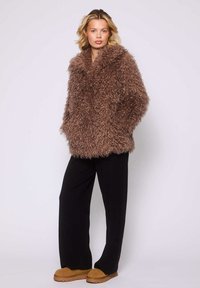 Cappotto in pelliccia sintetica marrone e shaggy con un ampio colletto, maniche lunghe e vestibilità morbida. Abbinato a pantaloni neri e scarpe slip-on color beige.