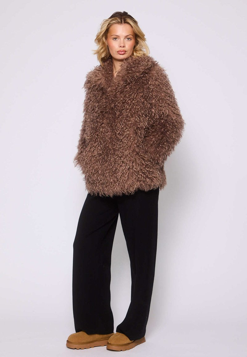 Cappotto in pelliccia sintetica marrone e shaggy con un ampio colletto, maniche lunghe e vestibilità morbida. Abbinato a pantaloni neri e scarpe slip-on color beige.
