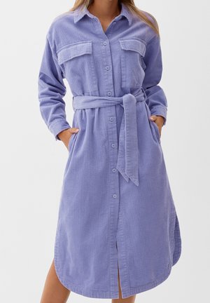 Kobieta ubrana w długą sukienkę koszulową z liliowego sztruksu z długim rękawem, kieszeniami na piersi i paskiem wiązanym w talii, z rękami w bocznych kieszeniach.