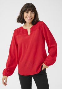 Röd långärmad blus med en texturerad, rynkad halsringning och en subtil lyster. Har en nyckelhålsöppning och en avslappnad passform. Kombineras med svarta byxor.