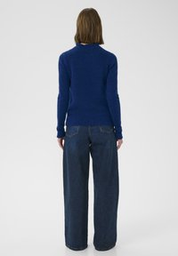 Modrý pletený sveter s vysokým golierom, dlhými rukávmi a priliehavým pásom. Kombinovaný s tmavomodrými džínsami so širokými nohavicami, ktoré majú textúrovaný povrch.