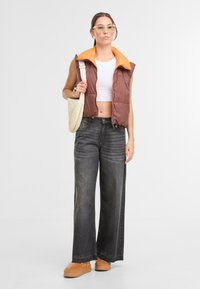 Hnedá a oranžová nadýchaná bunda bez rukávov cez biely crop top, doplnená širokými džínsami tmavej denim farby a hnedými platformovými topánkami. Drží krémovú kabelku na rameno.