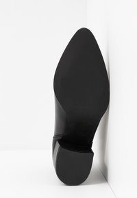 Semelle de chaussure habillée noire à bout pointu avec petit talon, positionnée verticalement contre un mur d'angle blanc.
