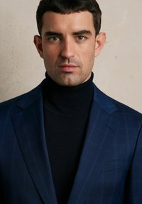 Granatowa marynarka w delikatny wzór w kratkę, zestawiona z czarnym golfem. Gładki materiał, usztywnione ramiona i starannie dopasowane detale.