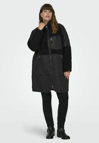 Black quilted long coat featuring plush upper, zippered pockets, high collar, and textured fabric. Paired with black slim-fitting pants and ankle boots.