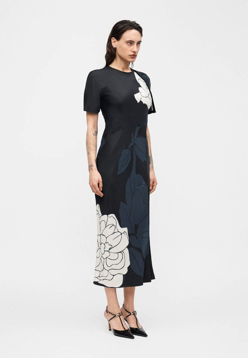 Femme portant une robe midi noire avec de grandes imprimés floraux blancs et foncés, manches courtes, et des escarpins noirs pointus avec des brides à la cheville.