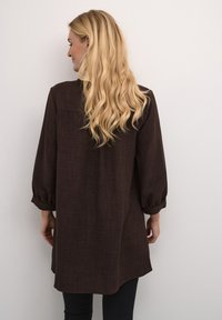Tunique marron en tissu texturé, à manches longues resserrées aux poignets, et coupe ample, vue de dos sur un fond uni.