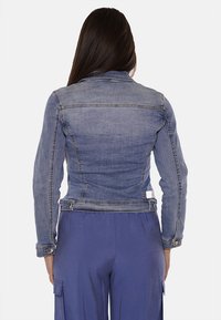 Giacca in denim di color blu chiaro, design aderente con maniche lunghe, cucitura della spalla posteriore e dettagli in metallo sui bottoni. Indossata sopra pantaloni viola.